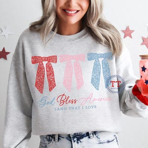 Puede incluir: Una sudadera gris con un dise&ntilde;o de lazos rojo, rosa y azul. El texto "God Bless America Land That I Love" est&aacute; impreso debajo de los lazos.