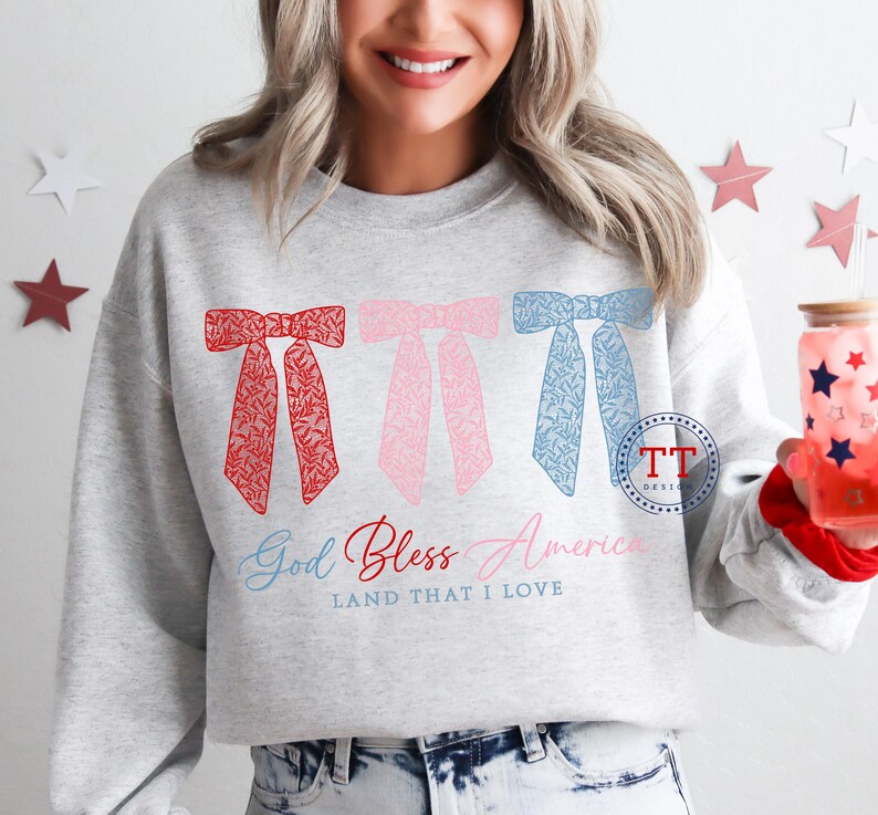 Puede incluir: Una sudadera gris con un dise&ntilde;o de lazos rojo, rosa y azul. El texto "God Bless America Land That I Love" est&aacute; impreso debajo de los lazos.