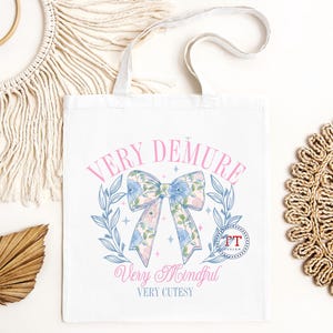 Peut inclure: Sac fourre-tout blanc avec un n&oelig;ud floral rose et bleu. Le texte "Very Demure" est &eacute;crit en rose au-dessus du n&oelig;ud et "Very Mindful" et "Very Cutesy" sont &eacute;crits en rose en dessous du n&oelig;ud.