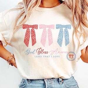 Puede incluir: Una camiseta blanca con un dise&ntilde;o de lazos rojo, rosa y azul. El texto "God Bless America LAND THAT I LOVE" est&aacute; impreso debajo de los lazos. Un peque&ntilde;o c&iacute;rculo con el texto "TT DESIGN" est&aacute; en la esquina inferior derecha.