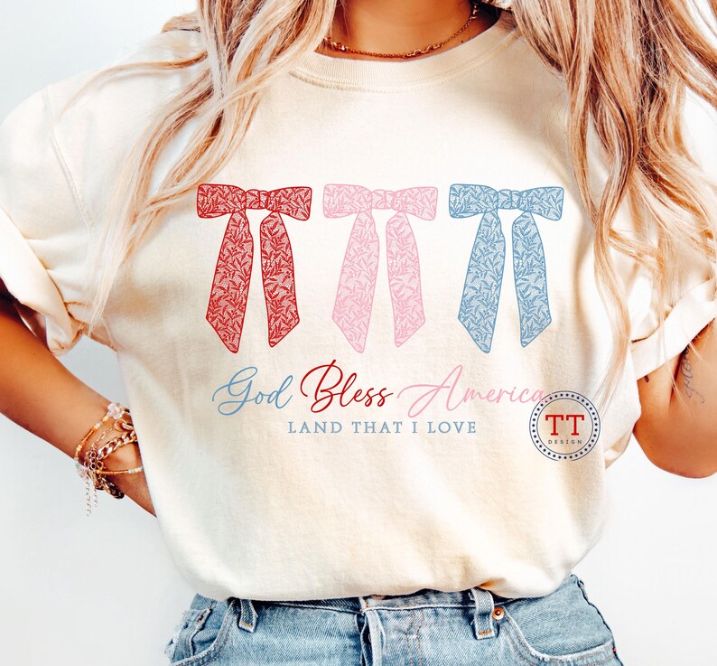 Puede incluir: Una camiseta blanca con un dise&ntilde;o de lazos rojo, rosa y azul. El texto "God Bless America LAND THAT I LOVE" est&aacute; impreso debajo de los lazos. Un peque&ntilde;o c&iacute;rculo con el texto "TT DESIGN" est&aacute; en la esquina inferior derecha.