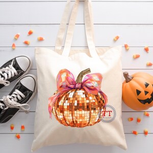 K&ouml;nnte beinhalten: Eine wei&szlig;e Canvas-Tragetasche mit einem goldenen Glitzerk&uuml;rbis mit einer rosa Schleife. Der K&uuml;rbis hat ein Discokugel-Design. Die Tasche ist perfekt zum Tragen von Halloween-S&uuml;&szlig;igkeiten.