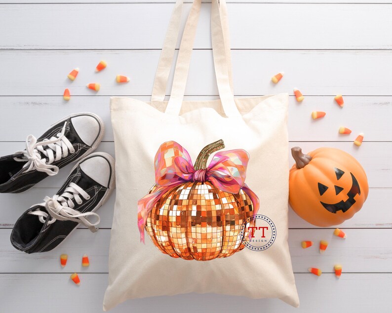 K&ouml;nnte beinhalten: Eine wei&szlig;e Canvas-Tragetasche mit einem goldenen Glitzerk&uuml;rbis mit einer rosa Schleife. Der K&uuml;rbis hat ein Discokugel-Design. Die Tasche ist perfekt zum Tragen von Halloween-S&uuml;&szlig;igkeiten.