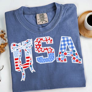 May include: Blue Comfort Colors t-shirt with the word "USA" in a patchwork design. The "U" is a bow with red and white stripes and blue patterns. The "S" is red with white floral accents, and the "A" is blue gingham and floral.