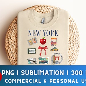 Op de afbeelding: Een beige sweatshirt met een grafisch ontwerp met de tekst "NEW YORK" in blauwe letters en verschillende illustraties van bezienswaardigheden in New York City, waaronder een rode appel, een gele taxi en een hotdogkraam.