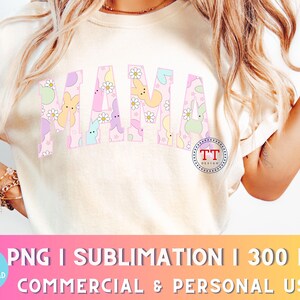 Puede incluir: Una camiseta blanca con un dise&ntilde;o gr&aacute;fico rosa, azul y amarillo que dice "MAMA" en letras grandes. El gr&aacute;fico presenta conejos y flores de colores pastel.