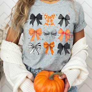 Pu&ograve; includere: Una maglietta grigia con un design di nove fiocchi diversi in nero, bianco, arancione e rosso. I fiocchi sono disposti in tre file di tre. La maglietta &egrave; indossata da una persona che tiene una zucca.