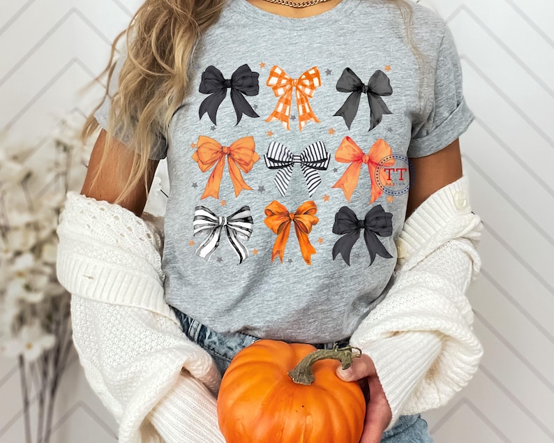 Pu&ograve; includere: Una maglietta grigia con un design di nove fiocchi diversi in nero, bianco, arancione e rosso. I fiocchi sono disposti in tre file di tre. La maglietta &egrave; indossata da una persona che tiene una zucca.