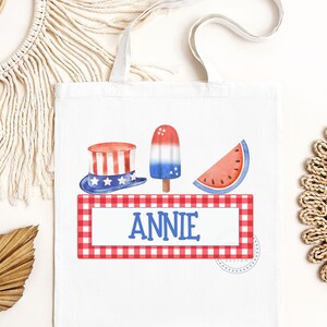 May include: A white tote bag with a red, white, and blue gingham border. The bag features a red, white, and blue popsicle, a watermelon slice, and a red, white, and blue top hat. The name "ANNIE" is printed in blue on the bag.
