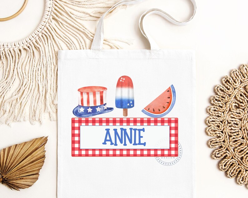 May include: A white tote bag with a red, white, and blue gingham border. The bag features a red, white, and blue popsicle, a watermelon slice, and a red, white, and blue top hat. The name "ANNIE" is printed in blue on the bag.