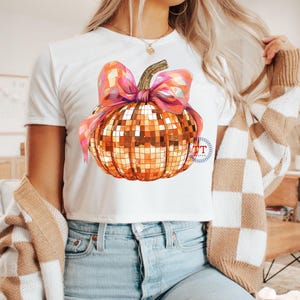 K&ouml;nnte beinhalten: Ein wei&szlig;es T-Shirt mit einem goldenen und rosafarbenen Glitzerk&uuml;rbis-Design. Der K&uuml;rbis hat eine rosafarbene Schleife oben.