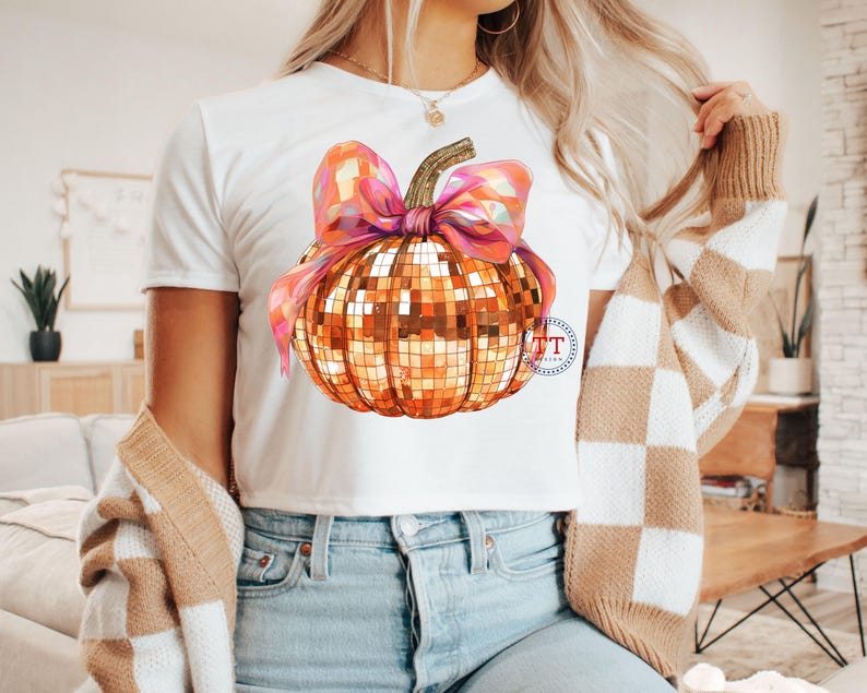 K&ouml;nnte beinhalten: Ein wei&szlig;es T-Shirt mit einem goldenen und rosafarbenen Glitzerk&uuml;rbis-Design. Der K&uuml;rbis hat eine rosafarbene Schleife oben.