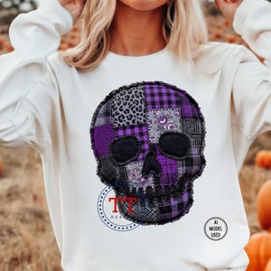 Op de afbeelding: Een crèmekleurig sweatshirt met lange mouwen en een grote, centrale afbeelding van een schedel die is samengesteld uit verschillende paarse, zwarte en grijze patchworkpatronen, waaronder luipaardprint, ruiten en bloemmotieven. De tekst TT DESIGNS is zichtbaar.