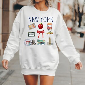 Op de afbeelding: Witte trui met een blauwe "NEW YORK"-afbeelding en verschillende illustraties met het thema New York City, waaronder een rode appel, een gele taxi en de Brooklyn Bridge.