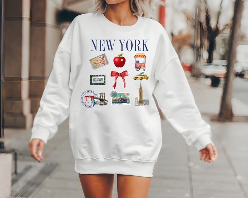 Op de afbeelding: Witte trui met een blauwe "NEW YORK"-afbeelding en verschillende illustraties met het thema New York City, waaronder een rode appel, een gele taxi en de Brooklyn Bridge.