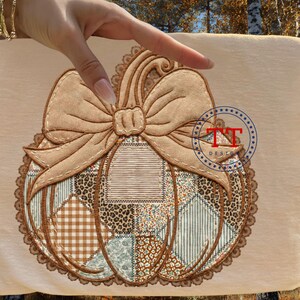 May include: A beige fabric with a detailed pumpkin design. The pumpkin has a patchwork pattern with leopard, plaid, and floral prints. A large bow sits on top, and the design is framed with a scalloped edge. The design has the text "TT DESIGNS".