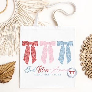 Puede incluir: Una bolsa de tela blanca con lazos rojos, rosas y azules con un patr&oacute;n floral. El texto "God Bless America Land That I Love" est&aacute; impreso en la bolsa.