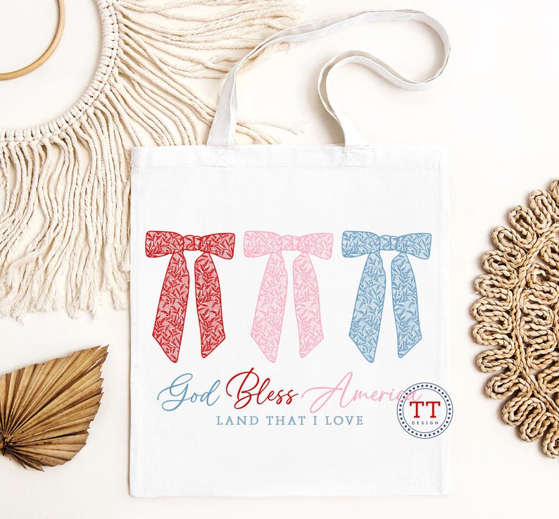Puede incluir: Una bolsa de tela blanca con lazos rojos, rosas y azules con un patr&oacute;n floral. El texto "God Bless America Land That I Love" est&aacute; impreso en la bolsa.