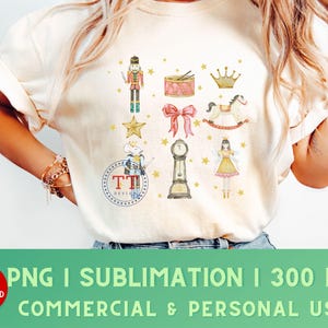 May include: Cream-colored t-shirt featuring a collection of watercolor illustrations. The design includes a nutcracker, drum, crown, bow, rocking horse, clock, fairy, and stars. The shirt also has the text "TT DESIGNS".
