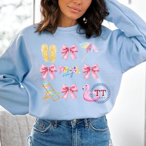 May include: Light blue sweatshirt with a summer theme design. The design includes pink bows, flip flops, a beach chair, a flamingo, an umbrella, and the words "Pool Party".