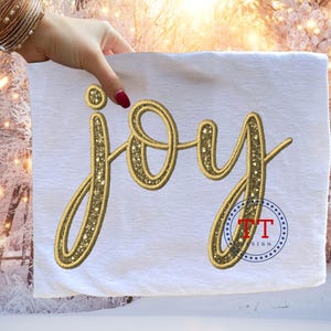 Könnte beinhalten: Weißer Stoff mit dem Wort "joy" in Goldfaden gestickt. Das Wort in Schreibschrift ist mit kleinen goldenen Sternen verziert. Ein kleines Logo ist in der unteren rechten Ecke sichtbar.