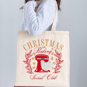 May include: A white canvas tote bag with the text "Christmas Baking Social Club" in gold lettering. A red stand mixer with a white bowl and a cup of coffee is in the center of the bag. The design is surrounded by a wreath of green leaves and brown gingerbread cookies.