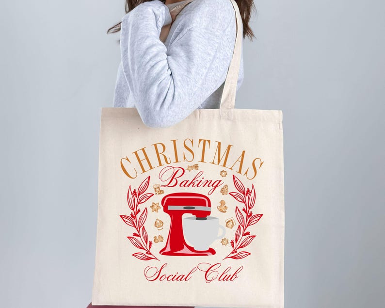 May include: A white canvas tote bag with the text "Christmas Baking Social Club" in gold lettering. A red stand mixer with a white bowl and a cup of coffee is in the center of the bag. The design is surrounded by a wreath of green leaves and brown gingerbread cookies.