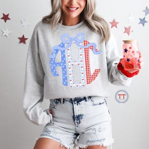 May include: A light gray sweatshirt with a large monogram design in red gingham, blue stars, and a white pattern. The monogram is decorated with a blue bow and red ribbon. The person is holding a glass with red and blue stars.