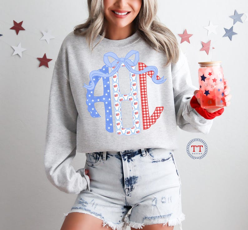 May include: A light gray sweatshirt with a large monogram design in red gingham, blue stars, and a white pattern. The monogram is decorated with a blue bow and red ribbon. The person is holding a glass with red and blue stars.