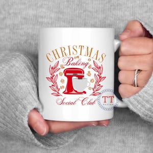 May include: White ceramic mug with a red and gold design. The design features a stand mixer, a wreath of holly, and the text "Christmas Baking Social Club".