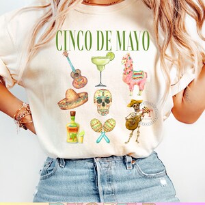 Pu&ograve; includere: Una maglietta bianca con il testo "Cinco de Mayo" in lettere verdi. La maglietta presenta una variet&agrave; di illustrazioni ad acquerello di icone culturali messicane, tra cui una chitarra, una margarita, un lama, un sombrero, uno scheletro, maracas e uno scheletro che suona la chitarra.
