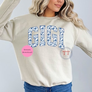 May include: A beige sweatshirt with the word "GIGI" printed in blue and white on the front. The letters are outlined in blue and filled with a blue and white floral pattern. The sweatshirt has a crew neck and long sleeves.