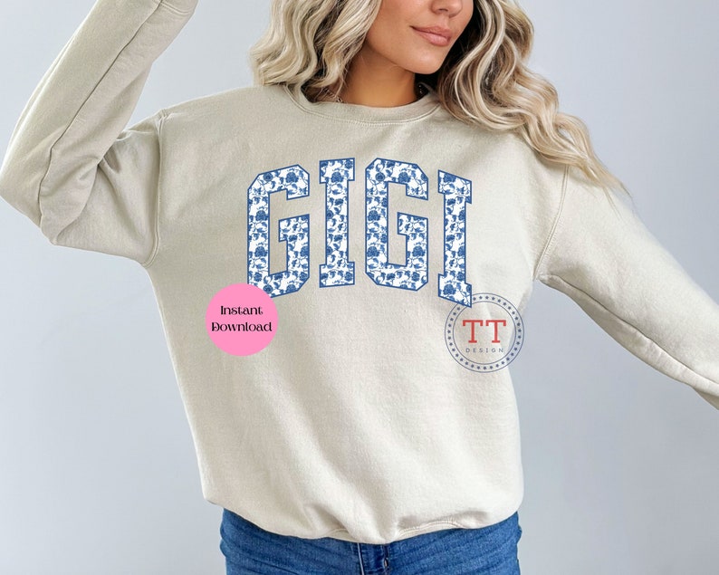 May include: A beige sweatshirt with the word "GIGI" printed in blue and white on the front. The letters are outlined in blue and filled with a blue and white floral pattern. The sweatshirt has a crew neck and long sleeves.