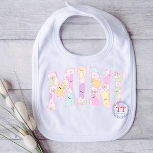 Puede incluir: Babero blanco con un patr&oacute;n pastel rosa, amarillo y azul de conejos y flores. El babero tiene la palabra "MINI" escrita en una fuente grande.
