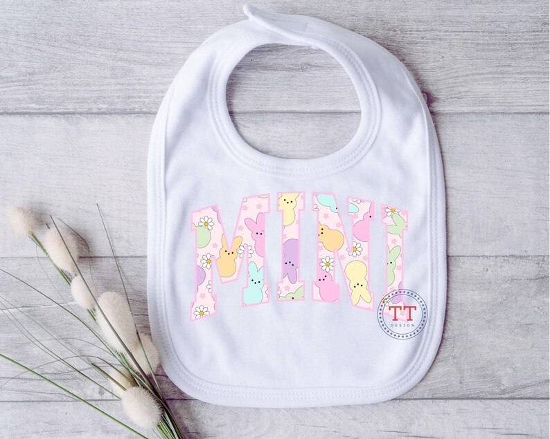 Puede incluir: Babero blanco con un patr&oacute;n pastel rosa, amarillo y azul de conejos y flores. El babero tiene la palabra "MINI" escrita en una fuente grande.