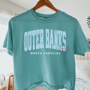 May include: A teal blue cropped t-shirt with the text "Outer Banks" in white with a blue outline and "North Carolina" in white below. The text "TT" is in red and white in a circle on the right side of the shirt.