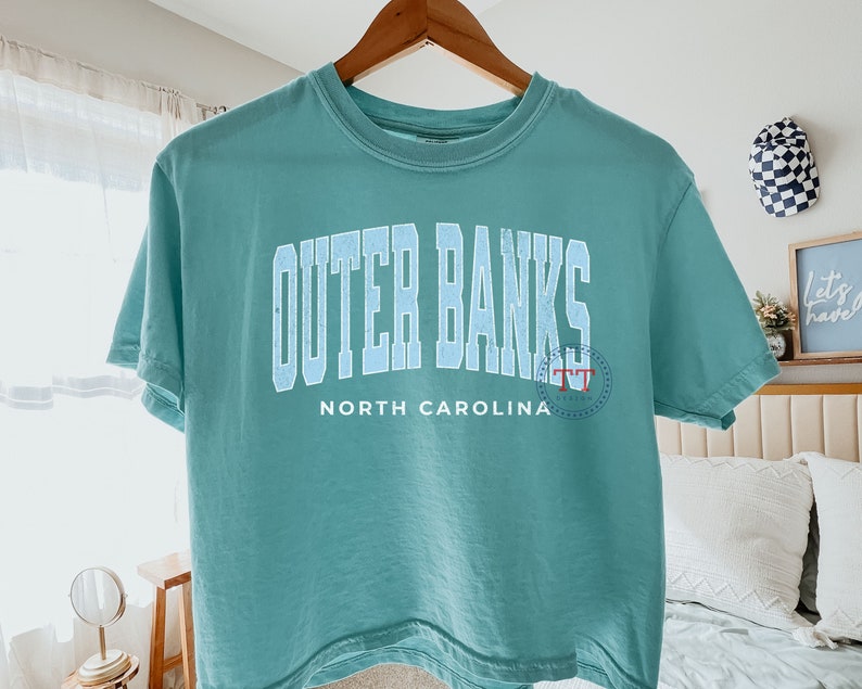 May include: A teal blue cropped t-shirt with the text "Outer Banks" in white with a blue outline and "North Carolina" in white below. The text "TT" is in red and white in a circle on the right side of the shirt.