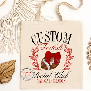 May include: A white canvas tote bag with black text that reads "CUSTOM Football Social Club TAILGATE SEASON". A red bow is on top of a brown football with red and green leaves around it. The text "TT DESIGN" is in a circle in the lower left corner.