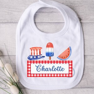 May include: White bib with a red, white, and blue checkered border. The bib features a red, white, and blue hat, a red, white, and blue popsicle, and a red watermelon slice. The text "Charlotte" is printed in blue on the bib.