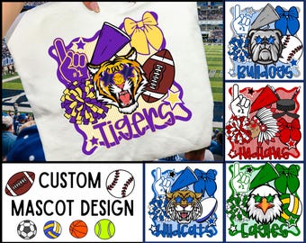 May include: A white t-shirt featuring a graphic print of a tiger mascot wearing star-shaped sunglasses, surrounded by a purple megaphone, a football, a yellow hair bow, and pom-poms, with the word Tigers written in purple script below.