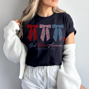 Puede incluir: Una camiseta negra con un dise&ntilde;o de lazo rojo, rosa y azul y el texto "God Bless America Land That I Love".