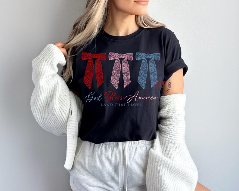 Puede incluir: Una camiseta negra con un dise&ntilde;o de lazo rojo, rosa y azul y el texto "God Bless America Land That I Love".