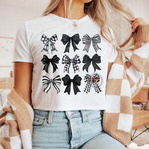 May include: White t-shirt with a black and white bow pattern. The bows are arranged in a grid pattern on the front of the shirt.