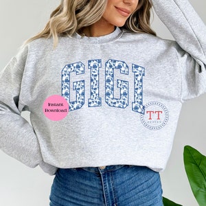 May include: A grey sweatshirt with the word "GIGI" printed in a blue and white floral pattern. The word is in a large, bold font. The sweatshirt is being worn by a person.