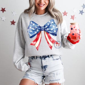 May include: A light gray sweatshirt featuring a large bow design in red, white, and blue, reminiscent of the American flag. The bow has a blue center with white stars and red and white striped ribbons. A glass with red stars is held in the hand.