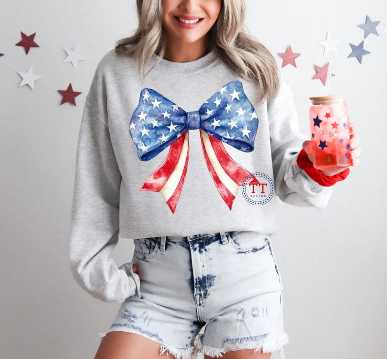 May include: A light gray sweatshirt featuring a large bow design in red, white, and blue, reminiscent of the American flag. The bow has a blue center with white stars and red and white striped ribbons. A glass with red stars is held in the hand.