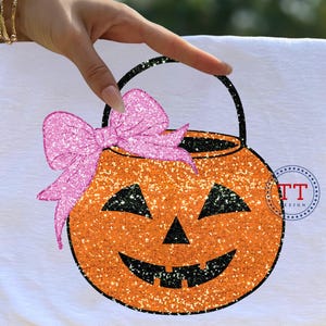 Puede incluir: Un diseño de Halloween con un cubo de calabaza naranja brillante con asa y cara negras. Un lazo rosa brillante está unido al cubo. El diseño está sobre un fondo blanco.