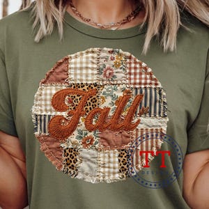 May include: Olive green t-shirt featuring a circular patchwork design with the word "Fall" embroidered in a warm brown color. The patchwork includes floral, plaid, and animal print patterns. A TT Designs logo is visible at the bottom right.