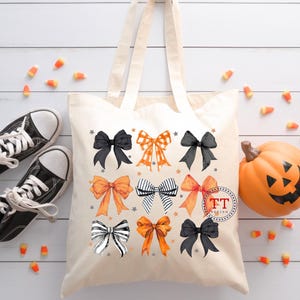 Pu&ograve; includere: Una borsa tote in tela bianca a tema Halloween. La borsa presenta diversi fiocchi arancioni, neri e bianchi con stelle. La borsa ha manici neri e il testo "TT Design" in un cerchio.