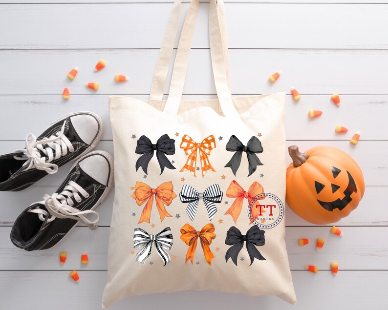 Pu&ograve; includere: Una borsa tote in tela bianca a tema Halloween. La borsa presenta diversi fiocchi arancioni, neri e bianchi con stelle. La borsa ha manici neri e il testo "TT Design" in un cerchio.